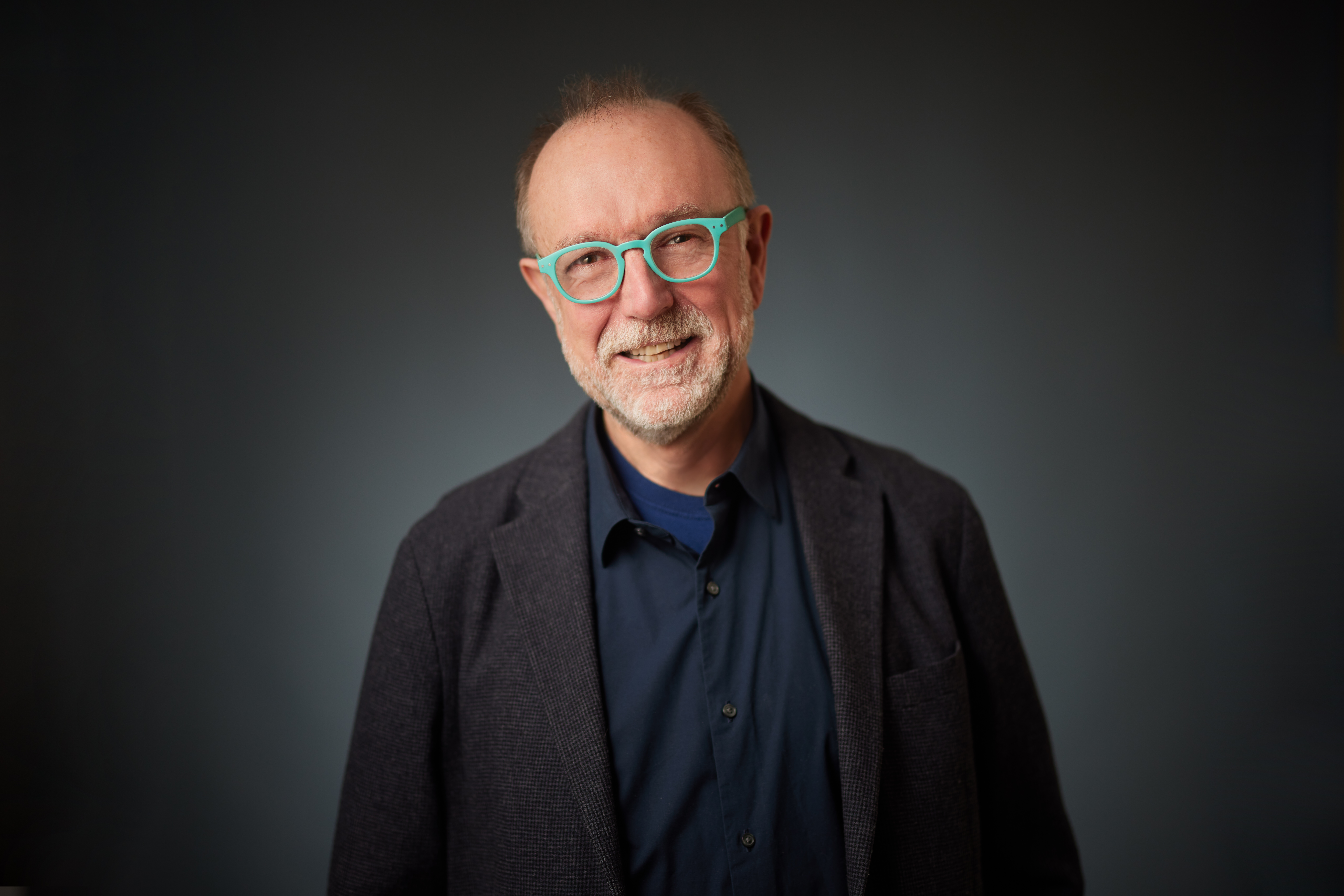 A headshot of professor Richard Clucas smiling wearing thick blue framed glasses.